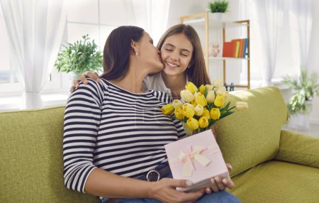 Photo for Happy child girl congratulating young mother with flowers tulips and a present box. Mom and daughter kissing and hugging sitting on the sofa at home. International Womens Day or Mothers Day. - Royalty Free Image