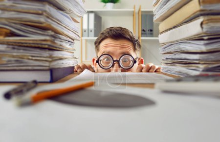 Photo for Overworked funny business man hiding under the desk at his workplace with a pile of folders and documents. Portrait of tired employee accountant working in office and looking at camera. - Royalty Free Image