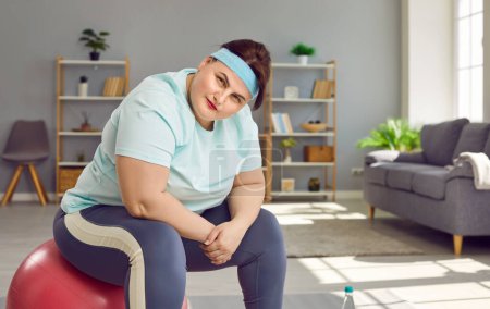 Photo for Portrait of a funny confident young fat woman wearing sportswear sitting on a fit ball at home and looking at the camera. Workout, sport exercises, fitness and body positive concept. - Royalty Free Image