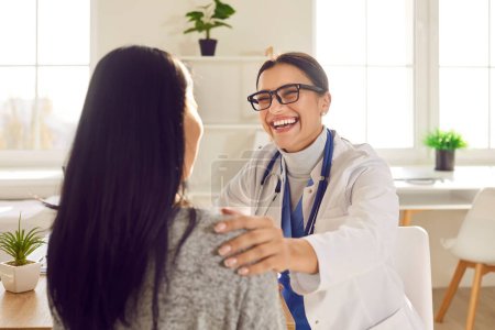 Téléchargez les photos : Joyeux docteur et patient qui s'amusent. Médecin femme en manteau médical blanc et lunettes parlant à la jeune femme, la réconfortant, lui tenant la main sur l'épaule, souriant, racontant une blague drôle et riant - en image libre de droit