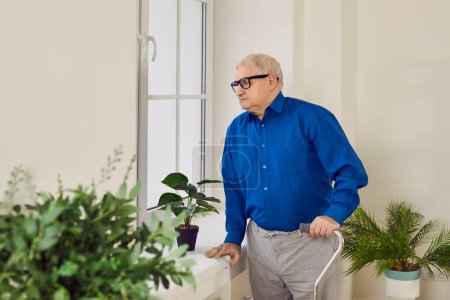 Portrait d'un aîné réfléchi regardant par la fenêtre. Vieil homme solitaire à la retraite avec canne à pied debout près de la fenêtre à la maison. Vieillissement, solitude, retraite, concept de handicap