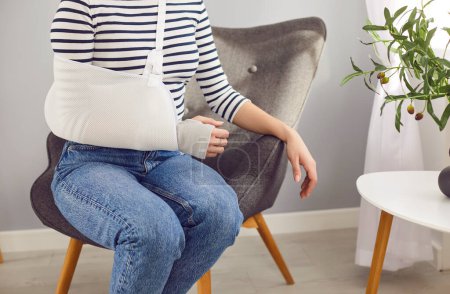 Crop unrecognizable young woman wearing a wrist bandage and an arm sling that help keep her injured limb immobile sitting on an armchair in the living room at home. Hand injury treatment concept
