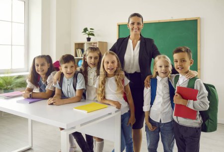 Erster Schultag. Klassenzimmer-Porträt einer Gruppe von Grundschülern und ihrer Lehrerin. Glückliche Kinder mit Rucksack und Notizbuch posieren mit ihrer Lehrerin und lächeln in die Kamera.