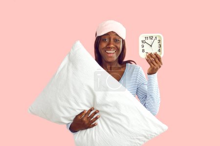 Téléchargez les photos : La jeune femme se réveille du sommeil le matin. Portrait de femme afro-américaine heureuse en pyjama debout isolé sur fond rose, tenant réveil et oreiller, regardant la caméra et souriant - en image libre de droit