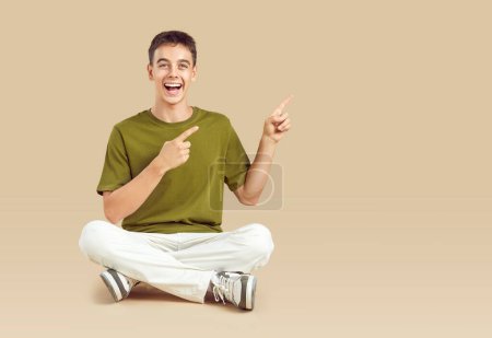 Photo for Cheerful teenager boy energetically points to advertising area on beige background. Funny Caucasian young man in casual clothes sits and laughs joyfully while pointing his fingers to side. Copy space. - Royalty Free Image