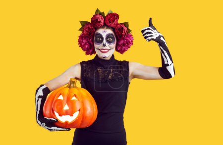 Photo for Studio portrait of happy funny woman with Catrina skull makeup standing isolated on yellow background, holding jack o lantern, smiling and showing thumbs up. Halloween, Day of Dead celebration concept - Royalty Free Image