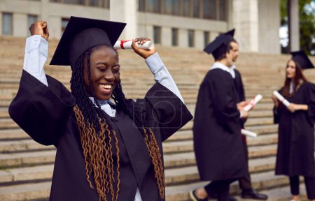 Foto de Joven estudiante afroamericana sonriente emocionada en un vestido universitario y diploma en sus manos. Chica graduada feliz haciendo sí gesto al aire libre en el fondo de compañeros de clase. Concepto de graduación. - Imagen libre de derechos