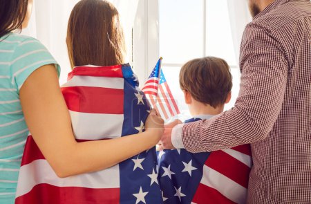 Téléchargez les photos : Vue arrière d'une jeune famille de quatre personnes avec des enfants garçon et fille debout à la fenêtre et regardant dehors à la maison enveloppé dans le drapeau des États-Unis célébrant le jour de l'indépendance. Patriotique US vacances. - en image libre de droit