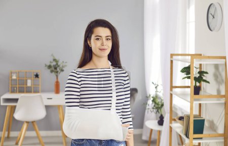 Photo for Smiling woman stands with bandage on her arm, showing strength and endurance despite injury. Portrait of smiling woman in stylish home interior wearing medical brace to support broken arm. - Royalty Free Image