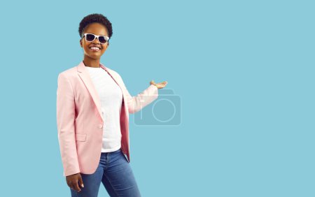 Téléchargez les photos : Femme afro-américaine souriante tenant la paume ouverte avec un espace de copie pour le produit publicitaire ou le texte au-dessus. Joyeux élégant jeune femme à la peau foncée présente un nouveau produit sur fond bleu clair. - en image libre de droit