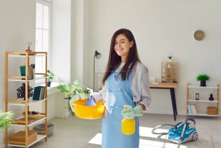 Photo for Happy housewife or maid service worker cleaning house. Beautiful smiling young woman in apron and rubber gloves holding work equipment and standing in living room, with vacuum cleaner in background - Royalty Free Image