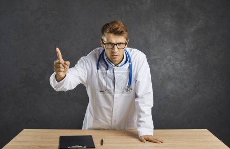 Photo for Angry doctor arguing and gesturing with his finger while standing at his desk on a gray background. Young Caucasian male doctor pointing his finger at you looking at the camera with a serious face. - Royalty Free Image