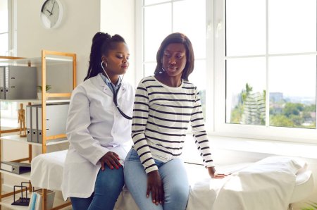 Téléchargez les photos : Une femme afro-américaine en blouse blanche examine une jeune patiente, lui pose un stéthoscope au dos et vérifie ses poumons et sa respiration. Bilan médical, médecine, concept de soins de santé - en image libre de droit