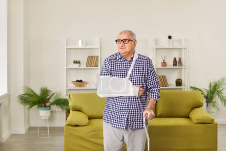 Photo for Injured senior man at home. Portrait of a retired, old man with a white cast and a sling on his broken arm standing in the living room and leaning on a walking stick. Injury concept - Royalty Free Image