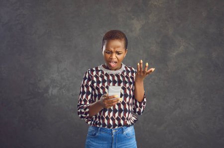 Photo for Angry African American woman waves her hand indignantly and shouts while looking at the screen of her phone. Woman reads an unpleasant message or is furious because of the lack of a mobile network. - Royalty Free Image