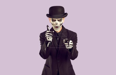 Photo for Aggressive Horror Movie Hero shoots at viewer with imaginary guns. Image of unkillable, ever-living mafia. Man in dark suit and black and white make-up. Isolate on light purple background. - Royalty Free Image