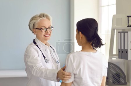 Doctor at the clinic or hospital supporting and reassuring her patient. Happy senior doctor in a white lab coat smiling and touching the young womans shoulder. Medicine and healthcare concept