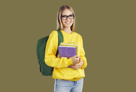 Lachende attraktive positive blonde Studentin in Brille mit Rucksack und Feilen auf khakifarbenem Hintergrund. Sie trägt Jeans und gelbes Sweatshirt. Bildung, Banner für Werbekonzept.