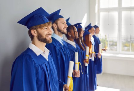 Photo for Portrait of a group of smiling happy multiracial diverse graduates students standing in a row in a blue university graduate gown and holding diploma indoors. Education and graduation concept. - Royalty Free Image