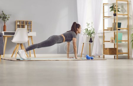 Foto de Retrato de una joven y atractiva mujer deportiva haciendo ejercicios de puja o deporte de tablón tumbada en una esterilla de yoga en el suelo de la sala de estar en casa. Fitness, entrenamiento y concepto de entrenamiento en casa. - Imagen libre de derechos