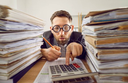 Business man in suit and funny glasses working at the desk on his workplace at office with a pile of folders and a stack of papers. Tired concentrated accountant making calculations analyzing company