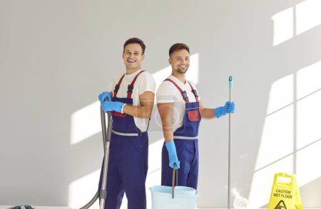 Photo for Team of happy male janitors from cleaning service. Two smiling men in overalls and rubber gloves, with mop, bucket and vacuum cleaner standing by light grey wall in room with caution wet floor sign - Royalty Free Image