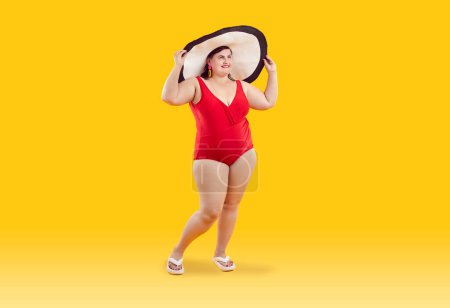 Photo for Portrait of cheerful female plus size model wearing beachwear, isolated on orange background. Chubby Caucasian woman posing in red one-piece swimsuit, beach flip flops and big straw hat. Full length. - Royalty Free Image
