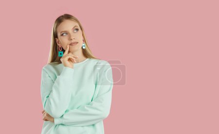 Photo for Thoughtful bewildered serious blondy girl in mint sweatshirt with geometric earrings looking up on copyspace on pink background. She touching her chin one hand. Banner for advertisement, marketing. - Royalty Free Image
