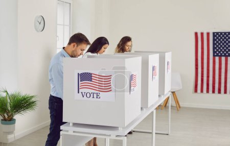 Téléchargez les photos : Les gens concentrés lisent l'information sur les bulletins de vote avant de voter. Des jeunes hommes et femmes votent anonymement dans les isoloirs d'un bureau de vote décoré de drapeaux américains. - en image libre de droit
