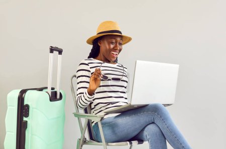 Photo for Excited surprised shocked happy smiling african american woman booking tickets for summer vacation online via laptop sitting on chair in beach hat with suitcase isolated on a studio gray background.. - Royalty Free Image