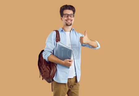 Portrait gutaussehender, lächelnder Student mit Brille, in lässiger Kleidung mit Rucksack auf dem Rücken, Notizbücher in der Hand und Daumen hoch. Isoliertes Studiofoto auf beigem Hintergrund.
