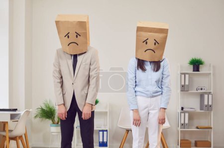 Foto de Dos compañeros de trabajo están juntos en la oficina llevando bolsas de papel en la cabeza, con emociones tristes y negativas. Las bolsas sirven como máscaras o disfraces, una experiencia compartida dentro del entorno empresarial. - Imagen libre de derechos