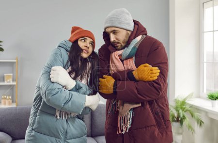 Warm gekleidete Männer und Frauen versuchen, sich zu Hause aufzuwärmen. Frustriertes Paar fühlt sich zu Hause kalt, während es im Wohnzimmer steht.Familienpaar in Winterkleidung versucht, Geld für ihre Stromrechnungen zu sparen