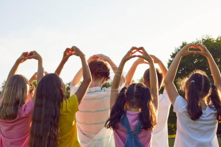 Téléchargez les photos : Vue arrière d'un groupe d'enfants faisant un geste en forme de cœur debout dans le parc d'été au coucher du soleil. Les enfants reconnaissants amis qui font signe amour levant la main dans la nature. Concept de protection de l'enfance - en image libre de droit