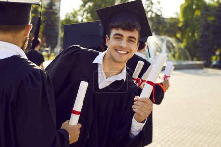 Photo for Portrait of happy smiling school, college or university student on graduation day. Young man in cap and gown with paper diploma scroll in hand looks back at camera while standing in line with others - Royalty Free Image
