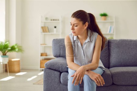 Photo for Portrait of upset sad depressed brunette woman sitting on the sofa in the living room at home alone. Young girl in bad thoughts feeling lonely indoors or having problems in relationships - Royalty Free Image