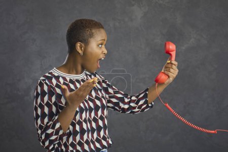 Photo for Wow. Excited black woman who got amazing news looking at red fixed land line cord phone receiver with open mouth and happy surprised face expression, side view studio portrait against grey background - Royalty Free Image