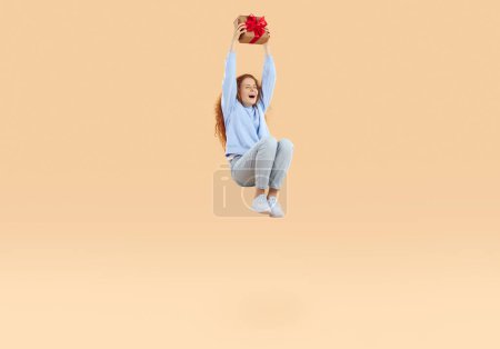 Glücklich lachende rothaarige lockige süße Mädchen in blauem Sweatshirt und Jeans hüpft in der Luft mit Weihnachtsgeschenkverpackung Bastelpapier mit roter Schleife, ein Geschenk auf beigem Hintergrund. Verkauf, Urlaubsrabatt.