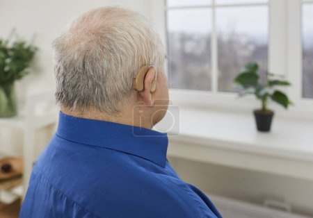 Foto de Acercamiento retrato de un hombre mayor de pelo gris sentado y usando audífonos modernos en su oído en casa mirando a la ventana con espacio para copiar. Concepto de pérdida auditiva y problemas de oído. - Imagen libre de derechos
