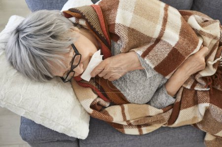 Téléchargez les photos : Gros plan portrait d'une femme âgée malade allongée sur un canapé à la maison enveloppée dans une couverture et souffrant d'écoulement nasal. Personne âgée de sexe féminin souffrant de grippe saisonnière se sentant en mauvaise santé avec la grippe. - en image libre de droit