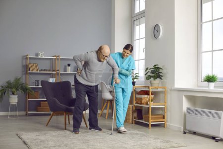 Téléchargez les photos : Une travailleuse médicale aide un aîné à marcher avec sa béquille. Joyeux soignant, infirmier en uniforme qui aide un homme âgé à se lever du fauteuil à l'intérieur. Soutien aux personnes âgées - en image libre de droit