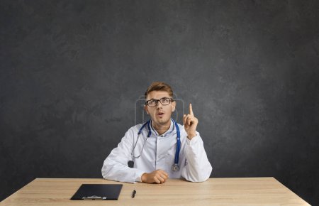 Pensive young male doctor in white medical uniform sit at desk hold finger up think of good idea or service. Man therapist look at blank empty copy space recommend medicine healthcare insurance.
