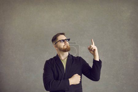 Photo for Funny guy thinks of a very smart idea. Young man in a suit jacket and thug life 8 bit meme glasses pointing his finger up isolated on a grey background - Royalty Free Image