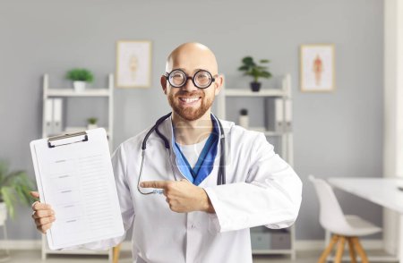 Photo for Portrait of young happy funny male doctor in white medical uniform standing in office pointing to report file with appointment and looking cheerful at camera in medical clinic. Health care concept. - Royalty Free Image