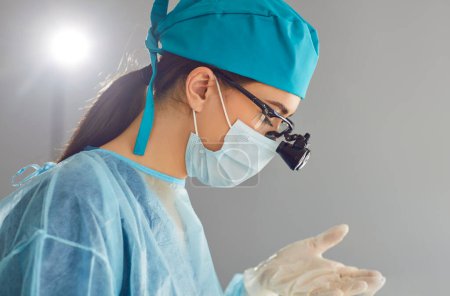 Nahaufnahme Porträt einer jungen Zahnärztin mit Binokularlupen im Gesicht, die eine blaue Maske und Uniform bei der Arbeit in der Zahnklinik trägt. Konzept zur zahnmedizinischen Versorgung.