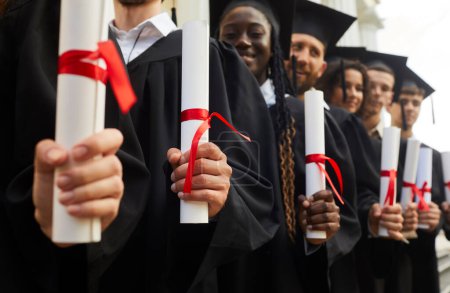 Photo for Happy confident graduate students standing in a row in black robes with diplomas in their hands outdoor at graduation ceremony. Education and graduating from university or college concept. - Royalty Free Image