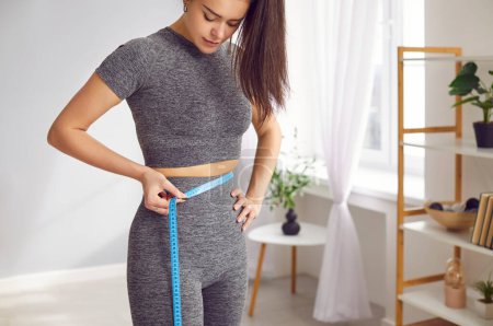Photo for Focused sportswoman checks her waist size with measuring tape, emphasizing importance of fitness. Young woman in stylish tracksuit measures her waist with measuring tape in her bright living room. - Royalty Free Image