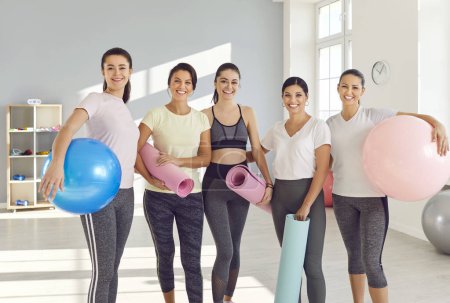 Téléchargez les photos : Portrait de joyeux groupe de femmes sportives gaies debout dans la salle de gym avec balle en forme et tapis de yoga dans les mains et regardant joyeusement la caméra après l'entraînement en forme. Entraînement sportif et mode de vie sain concept - en image libre de droit