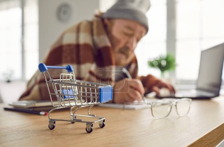 Photo for Close up of a small miniature shopping cart on a wooden table, with a poor senior retired man calculating and analyzing his expenses and planning his budget in the background. Budget concept - Royalty Free Image