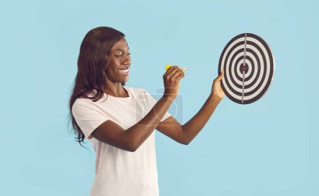 Téléchargez les photos : Portrait d'une jeune femme afro-américaine heureuse et joyeuse en vêtements décontractés visant flèche fléchette à la cible de tir avec expression de visage souriant isolé sur un fond de studio bleu. - en image libre de droit
